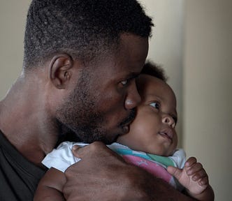 A Black father holding a baby inside a house, both looking in the same direction towards the right.