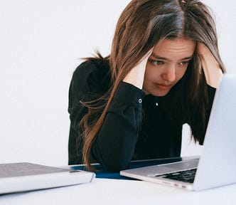 Woman wearning black shirt, holding her face in her hands, and looking at her computer screen. I suspect she works remotely and she just got an email that she is fired.