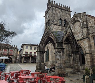 A medieval stone arch in front of a medieval church, with some red tables and chairs in front, under a dark cloudy sky.