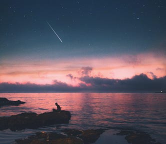 Shooting star over a pink sunset on a still body of water with a small human figure sitting in a boat to the lower left of the photo.
