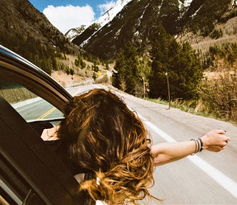 Back view of woman with long brown hair sticking head and arm out car window looking at road ahead