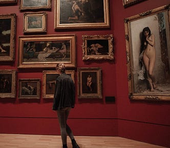 A young woman in an art gallery, admiring nineteenth century paintings. She has blonde hair worn up in a fishtail French braid, and she is wearing black ankle boots, grey leggings, and a blue denim jacket which is a little too large for her, so the sleeves cover her hands.