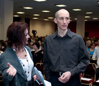 József Manhertz talks to the host of the event. The photo shows a lady on the left and a man on the right. The lady speaks looking towards the man and pointing her right palm upwards towards the camera. The man looks to the right while listening to the lady.