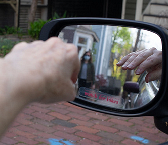 driver looks in rearview mirror at a pedestrian walking towards him wearing a mask and another person that is hard to see — the driver sees their own hand in the mirror very sharply.