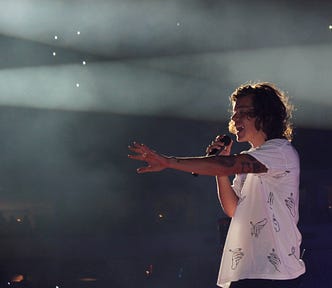 A young Harry Styles performing live, in a white shirt, in profile sideways shot from his left, with his right hand holding a microphone to his mouth and his left arm reaching out straight in front of him