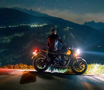 Man leaning against motorcycle at night as he stares into the distance over a town in the mountains