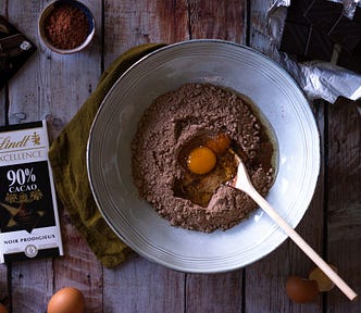 Metal bowl has a mix of flower and cocoa with a raw egg sitting on top and a wooden spoon. Around them on the wooden plank table are various bars of chocolate.