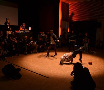 Dimly lit room with spotlight on a dance circle. Inside the dance circle is a breakdancer doing floor moves. An MC or the breakdancer’s opponent stands behind them. At the edge of the spotlight an audience of around 20 people.