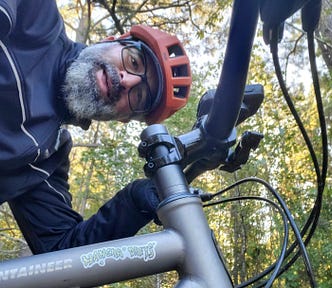 A person mountain-biking in a wooded area, wearing a safety helmet and cycling gear. Close-up view of a bicycle frame with the camera angle tilted, giving a dynamic feel