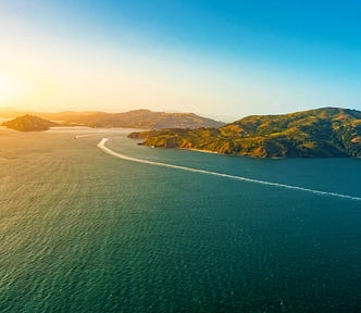 A view of Angel Island on the right side of the photo. The wake of two boats are visible in the strait. Rays of sunlight emanate from the left corner.