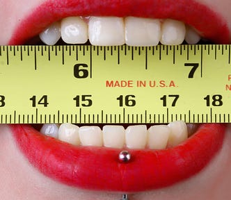 A very close up shot of a womans mouth with vibrant red lipstick, and a silver piercing in her lower lip- with a yellow measuring tape clenched between her teeth at the 6 inch mark.