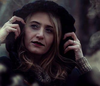 An attractive blonde woman in her forties wearing red lipstick looks mysterious in the woods while wearing a hood.
