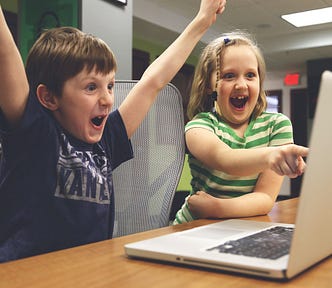 Children win! A young boy and Girl celebrate about what they see on a laptop. Boy puts 2 arms in the air saying Yay! and the girl points excitedly at something on the screen.