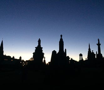 A photograph of a large cemetary (necropolis) in Glasgow. The sun is setting, leaving the sky a gradient of dark blue to white at the horizon. All the graves are in silhouette.