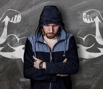 Young white man looking down, and crossing his arms against his chest, wearing a blue and grey hoodie and a serious expression, standing in front of muscular arms drawn on a chalkboard behind him.