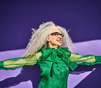 Portrait of a woman with gray curly hair and glasses wearing a green shirt against a purple background