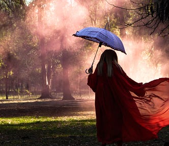 A woman wearing red, carrying an open blue umbrella approaching a patch of sunlight in the park.