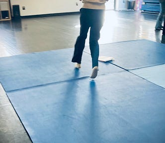 Image shows a pair of legs running through an open dance studio with a square of four blue foam yoga-like mats along the floor