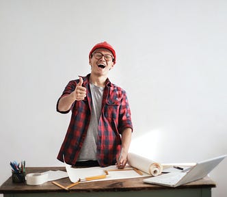 White man in a red shirt and red beanie grinning gormlessly at the camera with a thumbs up. He’s working on some sort of construction/architecture project; there are a lot of papers and pencils on his desk, as well as a laptop.