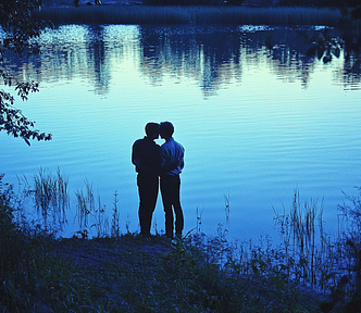 Two people standing close together by a serene lake at dusk, surrounded by foliage, with the water reflecting the twilight sky.
