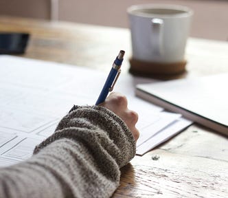 A close-up of a hand holding a blue pen, writing on printed documents spread across a rustic wooden desk, with a white ceramic mug on a cork coaster in the background.