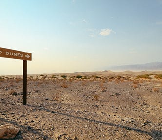An image from Unsplash showing a desert landscape with a single, somewhat forlorn road sign saying “Sand Dunes” with an arrow pointing toward the horizon.