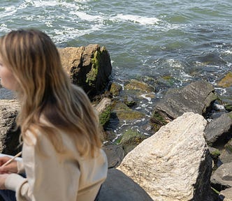 A woman is sitting on a rock next to a body of water, she has a pen and notebook and is writing while looking away from the camera