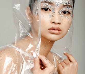 A woman of Asian descent dressed in a white singlet with white nail polish, holding a plastic hooded jacket. Her forehead is covered in silver glitter, as is her eye makeup. She is staring intensely at the camera with a grey backdrop.