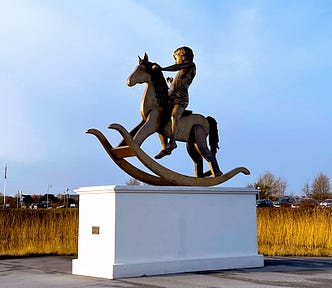 A larger-life bronze statue of a boy on a rocking horse, on a white plinth, background of winter-blue sky and a gold-brown bed of reeds