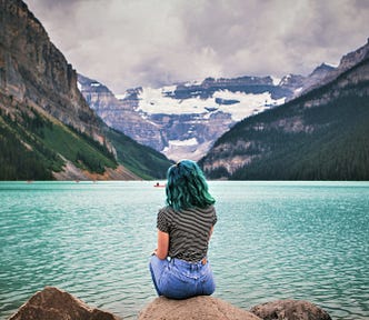 back view of girl wearing striped shirt and jeans sitting on rock looking out over lake and snowcapped mountains