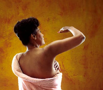 Back view of a black woman wrapped in a pink towel while doing a breast self-exam on the right side against an orange background.