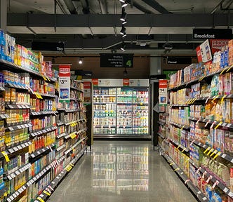 A well-lit grocery store aisle with shelves on both sides fully stocked with colorful boxes of cereal, tea, and breakfast items. Signs above indicate sections like “Tea,” “Cereal,” and “Breakfast.” Bright sale tags and price drop signs are visible on many shelves. At the end of the aisle is a refrigerated section filled with dairy products, and the polished floor reflects the lights and products. No people are present in the scene.