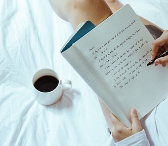 woman with coffee writing in notebook on bed