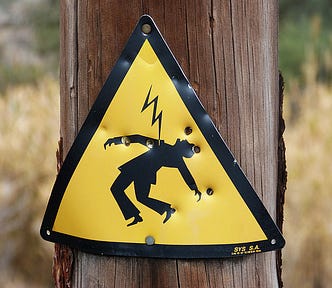A yellow, triangular ‘Danger of Death’ sign on a rural telephone pole, pockmarked with shotgun pellet holes.