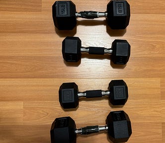 Four hexagonal dumbbells arranged in two columns on a wooden floor. The weights are labeled as 15 pounds and 10 pounds, with the 15’s  weight wrapped in tape on the handle for added grip. The dumbbells feature rubber-coated ends and metal handles with knurled grips.
