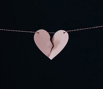 Image of a light pink, paper heart hanging from a thin red and white striped string. The paper heart is torn down the middle, from top almost to the bottom. The background is black.