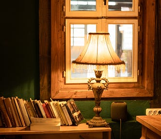 An antique light fixture with a cream coloured shade in front of a wooden framed window. The fixture sits on top of a book case that is jammed with a messy collection of books and papers.
