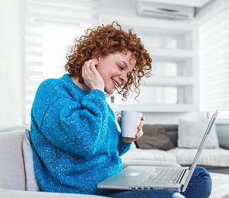 A happy woman in a blue sweater drinking coffee and writing on a laptop.