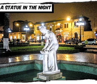 A white statue standing in a fountain in front of several buildings. It’s a night scene and lights are reflected on the slick pavement. The caption reads, “A statue in the night.”