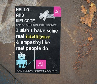 A sticker on a rusted metal background. The background of the sticker is black. The sticker shows a simple illustration of a small blue robot toy, the letters ‘AI’ in a pink square at the top and bottom, and the words, “Hello and welcome. Artificial intelligence. I wish I have some real intelligence and empathy like real people do. And funny? Forget about it.”