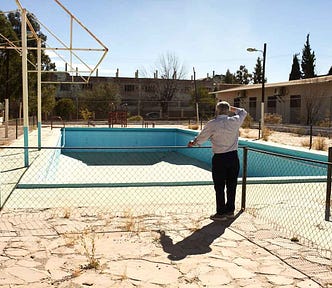 On a sunny day under a clear blue sky, an 80 year old man looks over an empty public swimming pool from behind a waist level chain link fence. The pool is large, painted bright blue and surrounding floor is made of flat polished stone. This man has not been here in almost 70 years. He is reminded of his childhood.