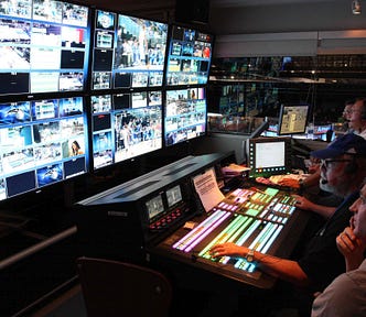 Several people sitting at a control room desk during a newscast, looking at several screens