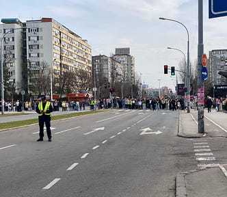 A singular police man blocks traffic while students stand across the intersection of a road.