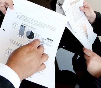 Two sets of hands going over a report. One is pointing at a graph and wearing a business suit, while the other set of hands is reading over the rest of the report.