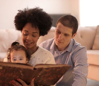 Interracial heterosexual couple of white male and brown-skinned female with short curly hair. The mom is holding a 2 year old on her lap and they’re reading her a story from a big, brown, old-looking book. They are sitting on a living room floor next to the couch.