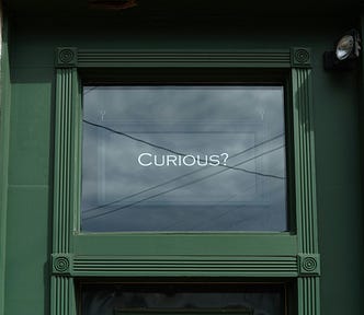 Top half of a green door with the word ‘curious?’ on the window