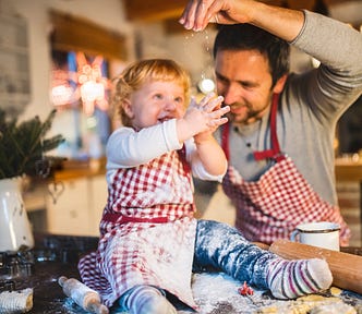 Father and his son make cookies and the father is sprinkling flour over sons hands to look like its snowing. The son plays with the falling flour in glee.