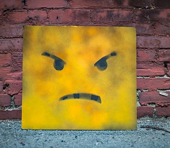 A canvas with a yellow angry face painted on it rests against a brick wall.