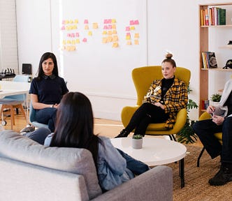 A relaxed meeting with five people sitting on comfortable chairs and on a sofa with post it notes on the wall