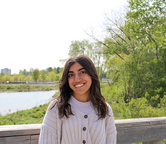 Headshot photo of Tara smiling at the camera. She has longer than shoulder length dark hair and is wearing a cream-colored cardigan. In the background, there is greenery and a lake.